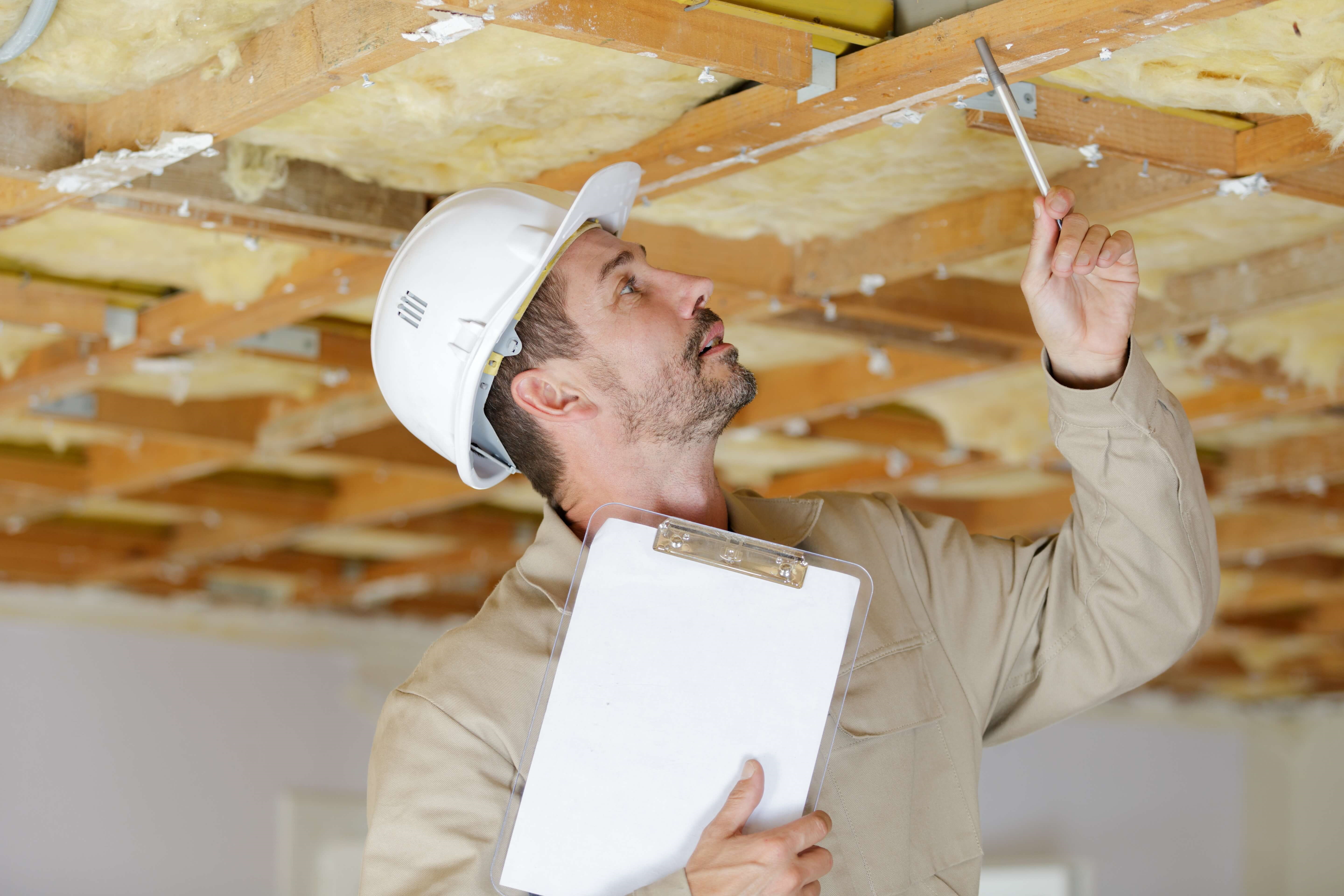 Mann in Bauleiter-Funktion mit Schutzhelm und Klemmbrett prüft die Decke eines Bauwerks auf Baumängel