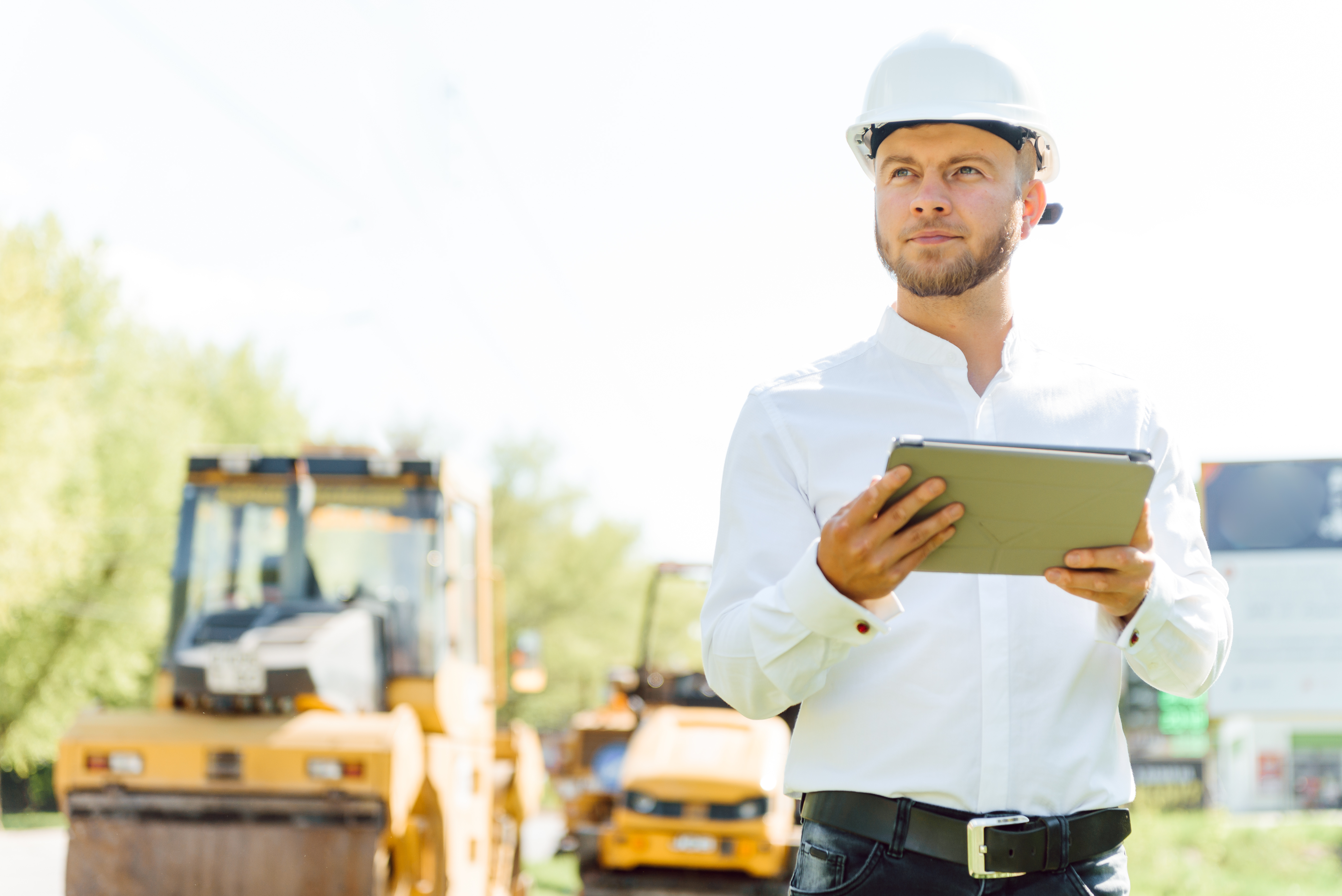 Mann in Hemd und mit Schutzhelm bei der Objektüberwachung mit Tablet in der Hand, im Hintergrund Baumaschinen