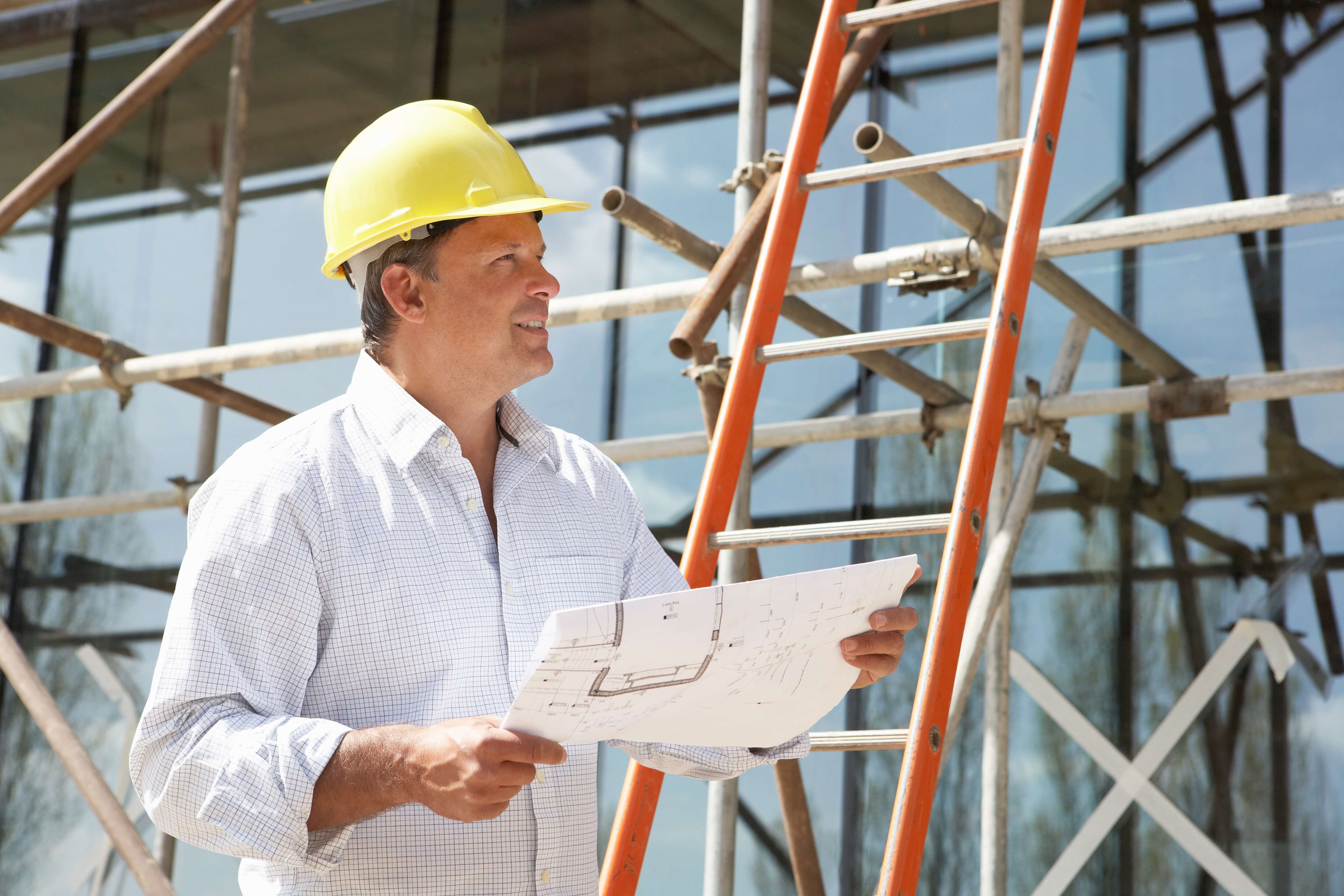 Mann mit Schutzhelm und Grundrisszeichnung steht auf einer Baustelle vor einer Leiter am Gerüst