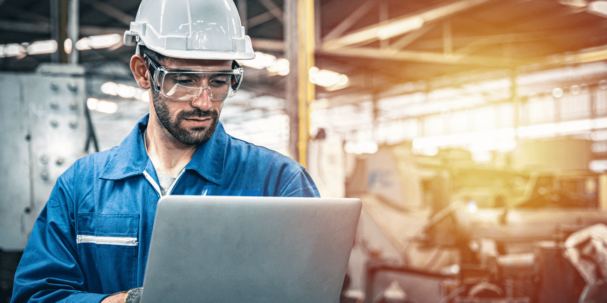 Mann in Blaumann und persönlicher Schutzausrüstung (Helm, Schutzbrille, Arbeitshandschuhe) in Fertigungshalle arbeitet an einem Laptop im Stehen