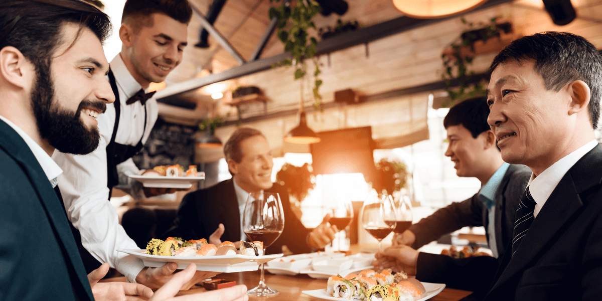 Geschäftsmänner mit kaukasischer und asiatischer Abstammung im Gespräch in einem Restaurant, ein Kellner serviert Sushi 