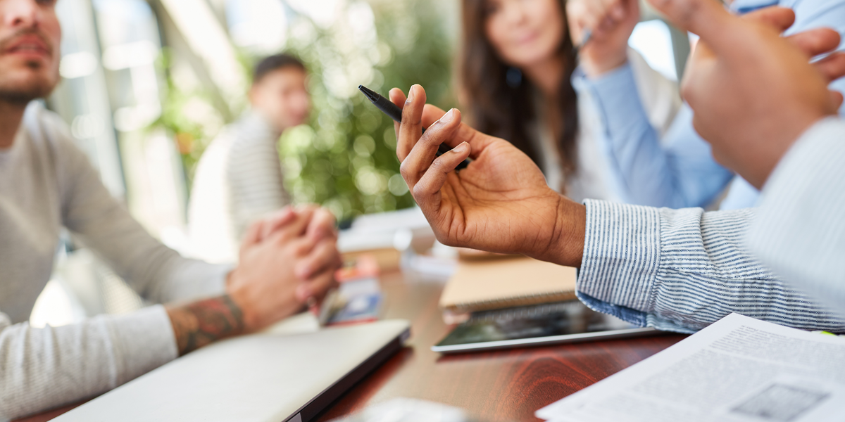 Junge Menschen diskutieren an einem Tisch mit Laptops, Tablets und Unterlagen, im Bildvordergrund gestikulieren Hände mit Stift 