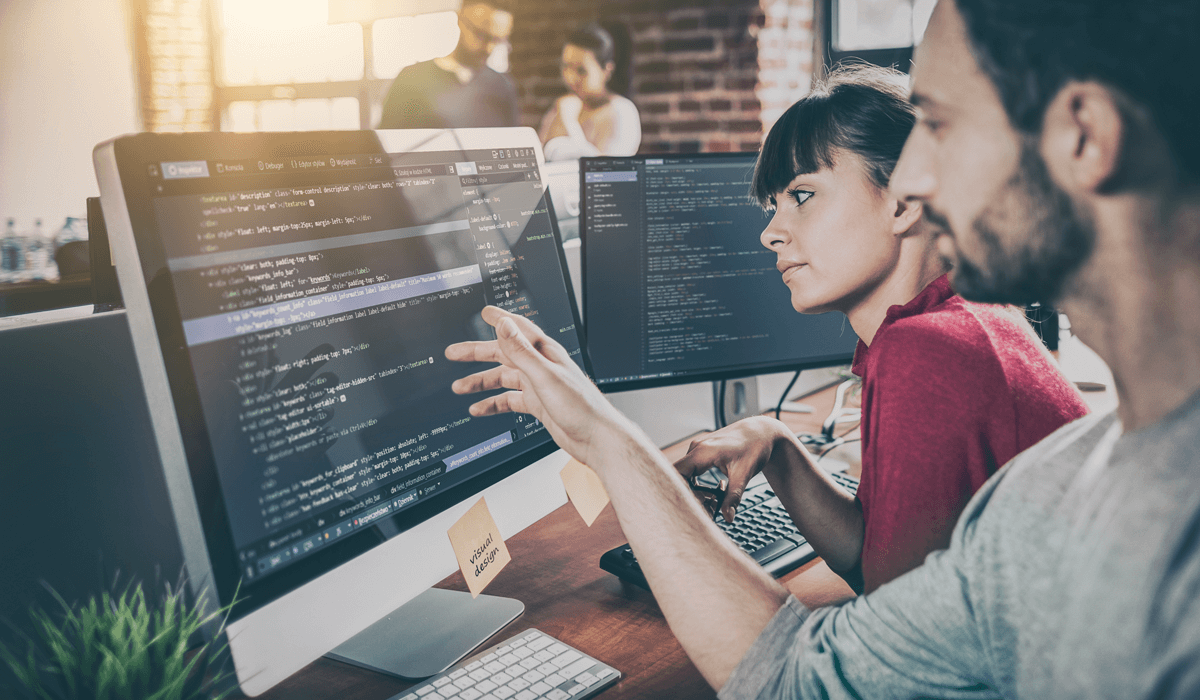 Zwei Kolleg/-innen überprüfen die technische Einbindung von Code auf einem Monitor in einem großen Büroraum 