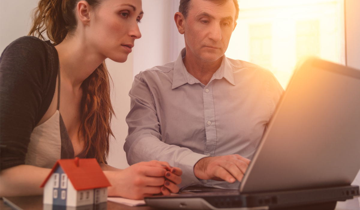 Zwei Personen blicken konzentriert auf einen Laptop-Bildschirm, auf dem Tisch steht ein Modellhaus 