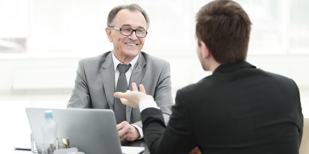 Älterer Mann in Business-Anzug an Meetingtisch mit Laptop lächelt im Gespräch mit einem jungen Mann