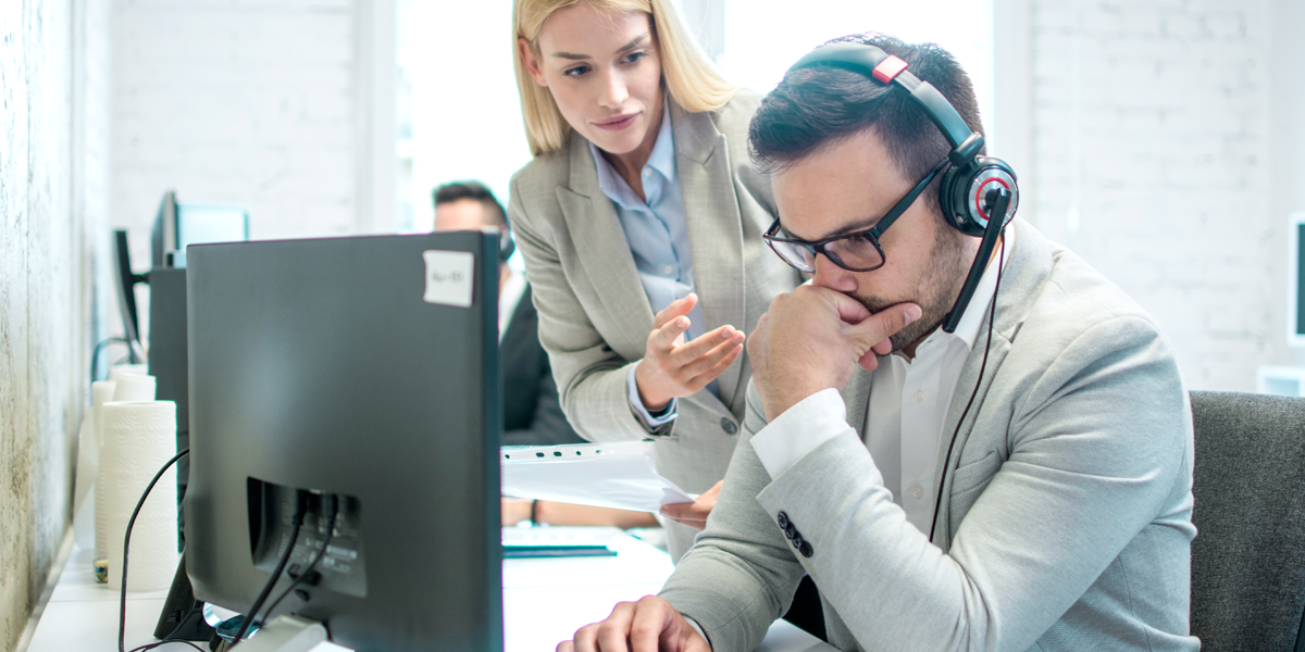 Mann in Anzug in nachdenklicher Haltung an Arbeitsplatz mit Working Station und Headset, eine Frau in Business-Outfit spricht ihn an