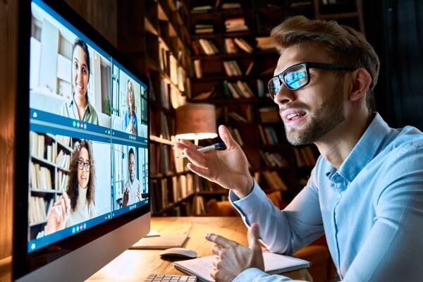 Junger Mann mit Brille vor dem Monitor in einer Arbeitsdiskussion per Video-Konferenz mit vier Mitarbeitenden