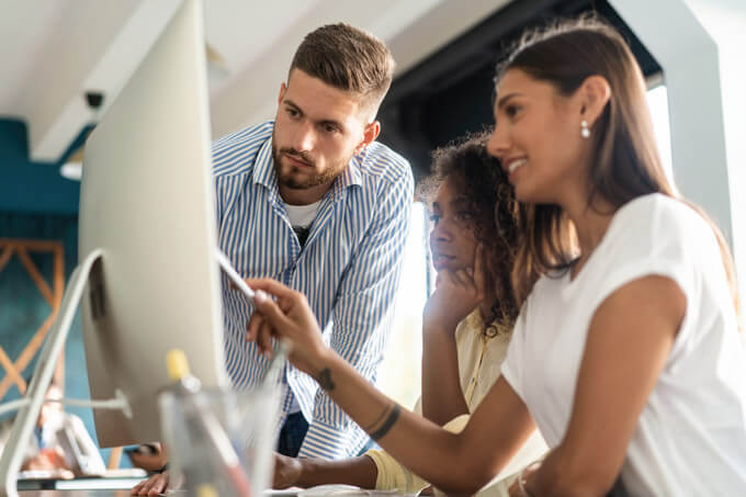 Junges Team von zwei Frauen und einem Mann schauen auf einen Bildschirm an einer Working Station