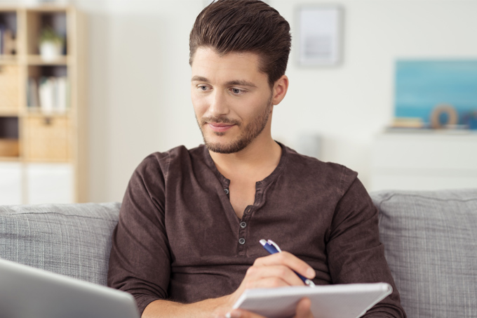 Mann sitzt auf dem Sofa, schaut interessiert auf den Laptop-Screen und macht auf einem Block handschriftliche Notizen
