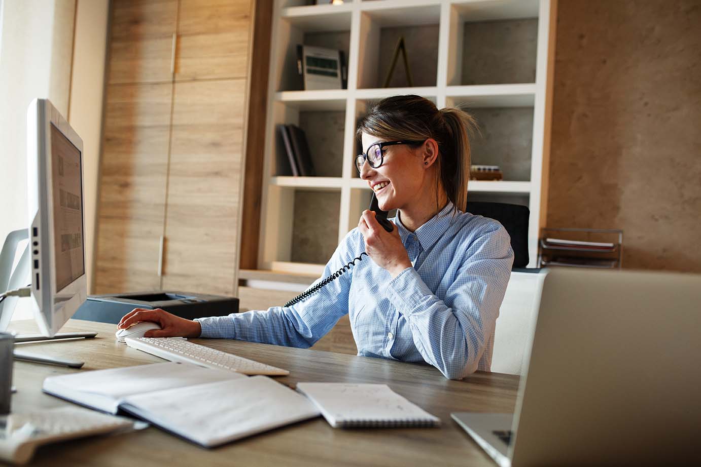 Geschäftsfrau an Schreibtisch arbeitet an Working Station am Schreibtisch, blickt auf den Monitor, telefoniert und lächelt