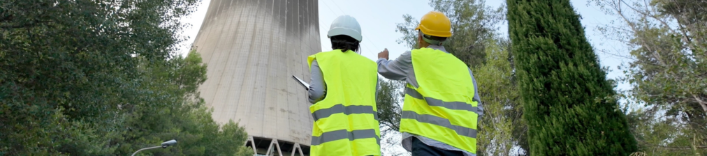 Zwei Fachkräfte in Warnwesten und Schutzhelmen inspizieren eine Industrieanlage mit Kühlturm.