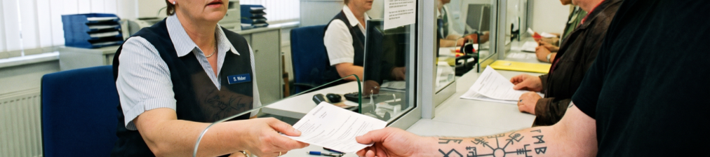 Mitarbeiterin an einem Serviceschalter übergibt einer Person mit tätowiertem Unterarm ein Dokument durch eine Glasscheibe.